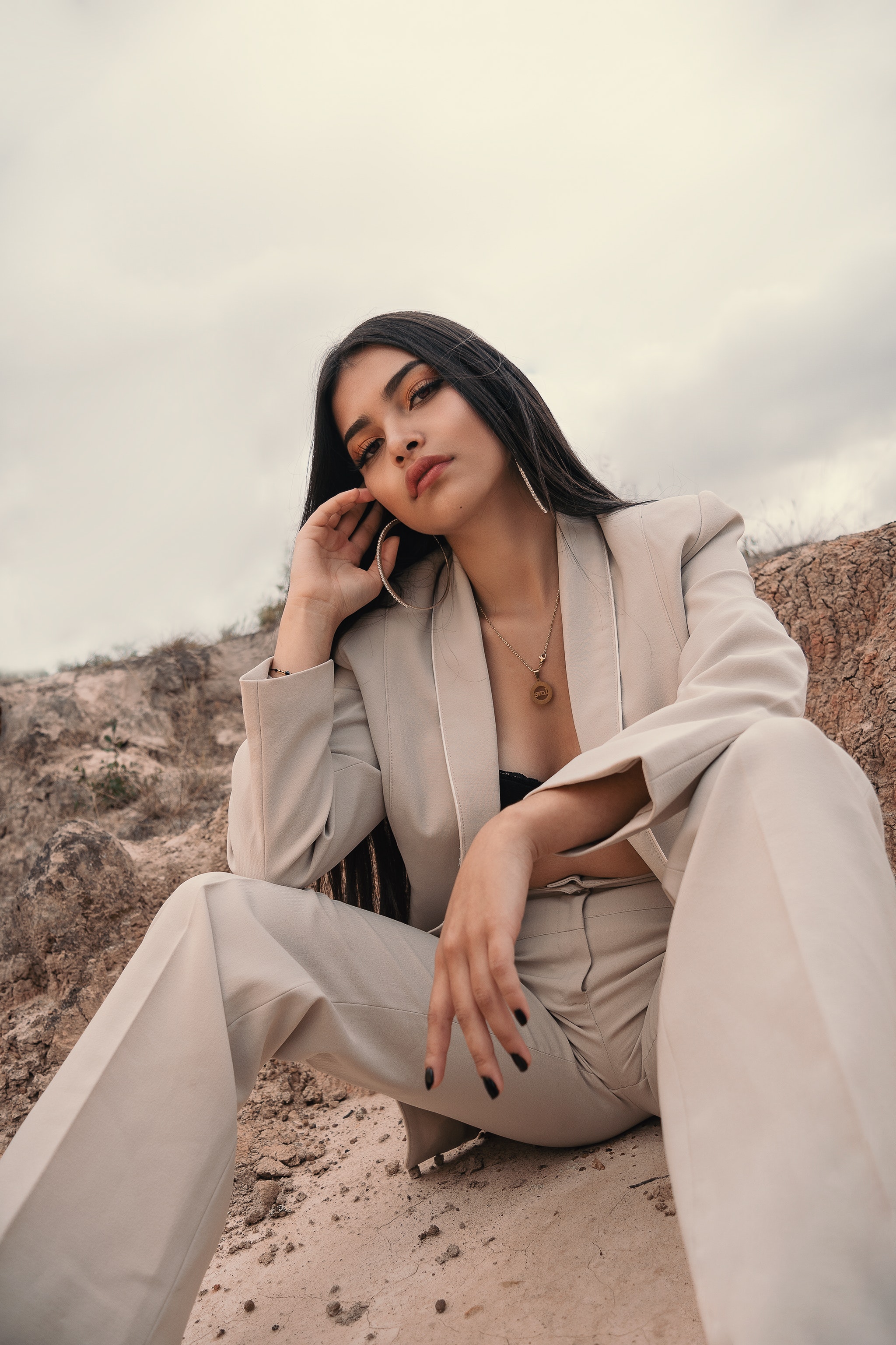 An elegant woman poses in the sand on the beach in a beige suit and dainty jewelry.