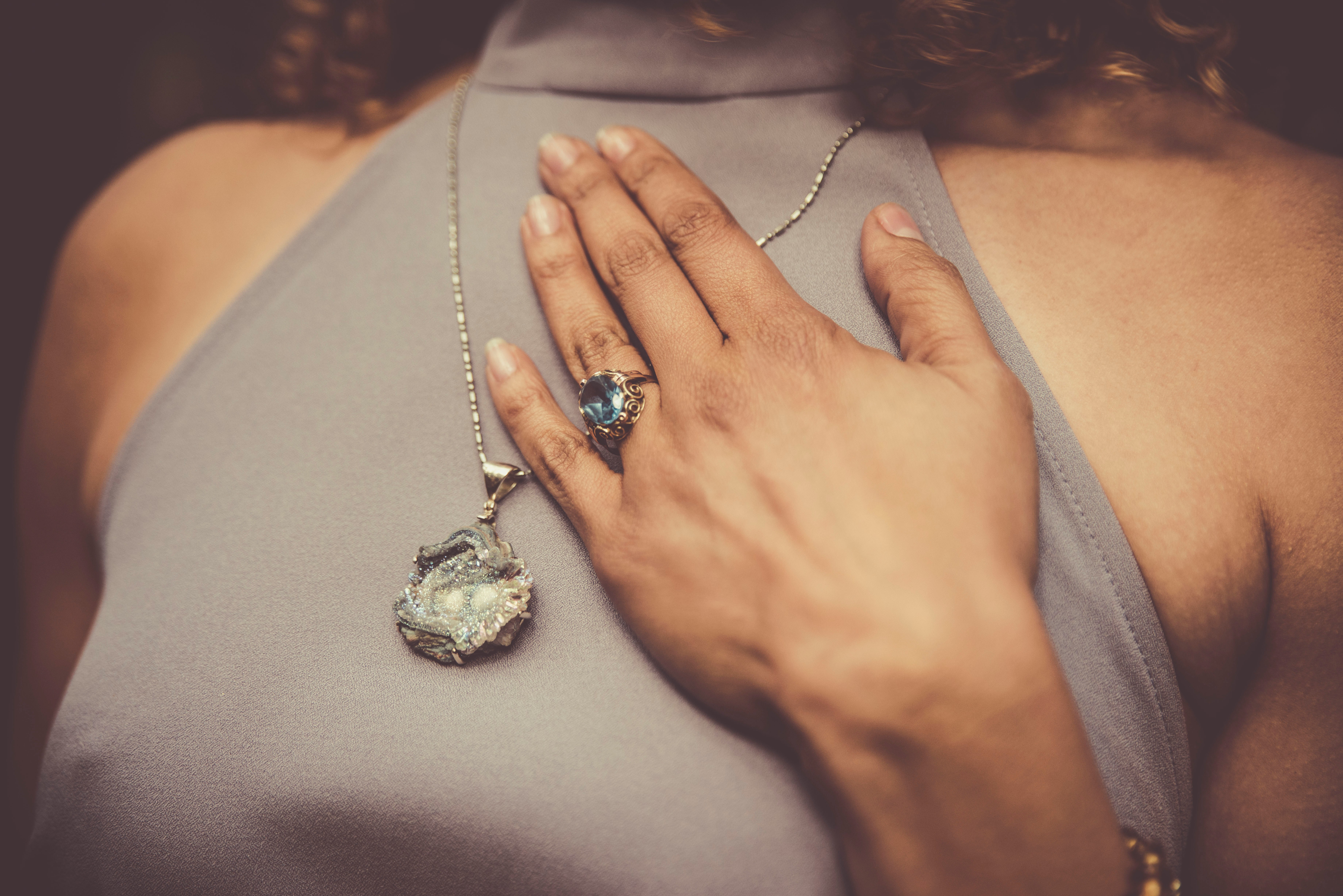 A woman from the neck down, showing off a big pendant necklace and an ornate aquamarine ring on the ring finger of her right hand.