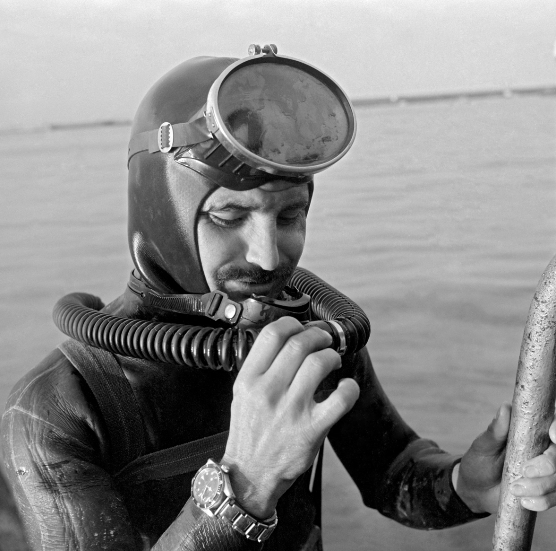 A black and white photo of a French diver emerging from the water in 1961, wearing his TUDOR submariner dive watch upside down on his right hand. The photo is sourced from the French Department of Defense Archives.